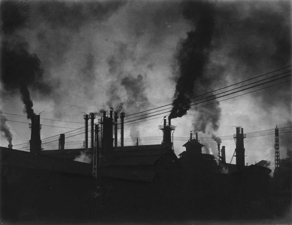 Photographie en noir et blanc représentant des cheminées d'usine dont sort de la fumée